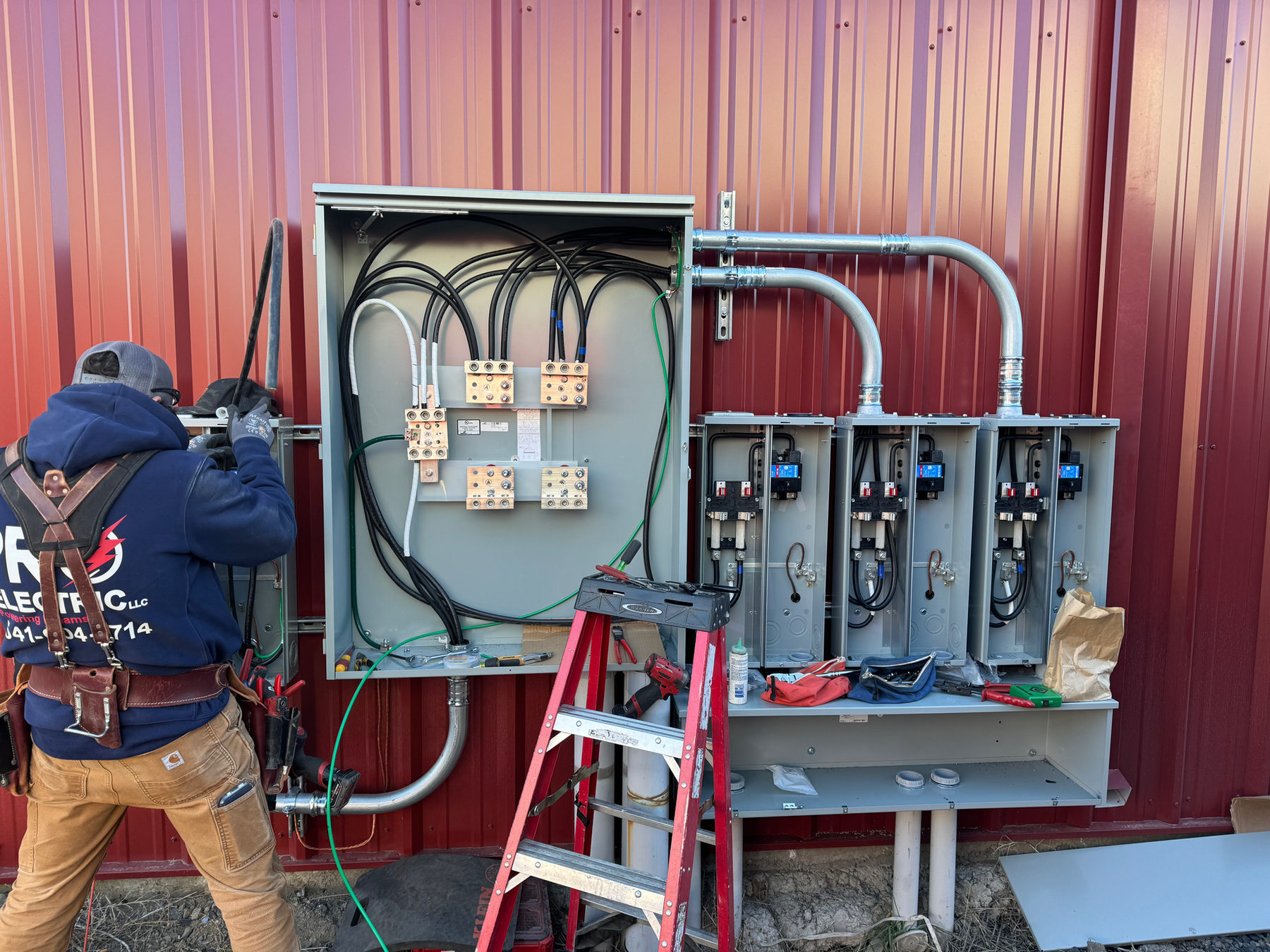 Pro Electric electrician wiring a multi-meter service bank on a red metal building